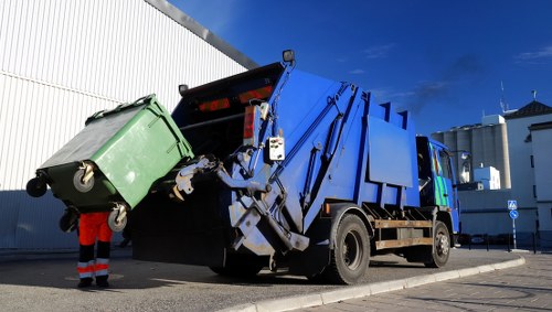 A large blue refuse collection vehicle with mechanical arms is actively lifting a green wheeled rubbish bin, which appears to be made of durable plastic with a textured surface, onto the back of the truck for waste removal. The truck is parked on a paved street with an industrial or commercial backdrop, including a white metal fence on the left and several white buildings with flat roofs and ventilation units in the background. The scene is set during daylight with a clear blue sky overhead. A worker wearing high-visibility clothing is partially visible on the left side of the image, assisting with the bin handling. This is typical of rubbish collection services provided by companies like House Clearance Brimsdown, covering areas including Brimsdown and surrounding postcode districts, and relates to the disposal or collection of household or commercial waste in the local area.