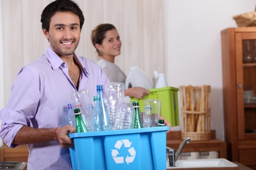 A young man with dark hair and a light purple shirt is smiling at the camera while holding a blue recycling bin filled with empty glass bottles and jars, including green and clear glass, situated in a domestic kitchen environment. Behind him, a woman with brown hair pulled back, wearing a light grey top, is turned slightly to the side and appears to be managing cardboard boxes and other household items on a countertop. The kitchen features wooden cabinetry, a stainless steel sink, and a light-colored curtain or wall covering in the background. The scene suggests a rubbish removal or house clearance activity, emphasizing recycling of glass bottles, in a typical UK home setting, consistent with the services offered by House Clearance Brimsdown near postcode EN8, in the vicinity of Brimsdown.
