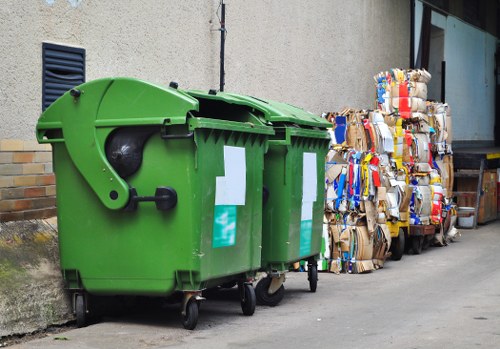 A large, bright green wheeled waste bin made of durable plastic with a partially open lid, positioned on a concrete pavement adjacent to a building wall. The bin has white waste collection stickers on its side and is situated in an outdoor urban setting. To the right of the bin, there are stacks of flattened cardboard boxes and packaging materials, with some cardboard pieces showing torn edges and creases, ready for collection or disposal. The background includes a grey stucco wall with a small black vent, suggesting a typical residential or commercial building environment, possibly within the Brimsdown area. The scene depicts typical rubbish removal elements, demonstrating the type of waste that a service like House Clearance Brimsdown handles, emphasizing waste collection and disposal in a professional context. The lighting is natural, indicating daytime conditions, and the overall setting reflects a typical urban street scene in the UK.